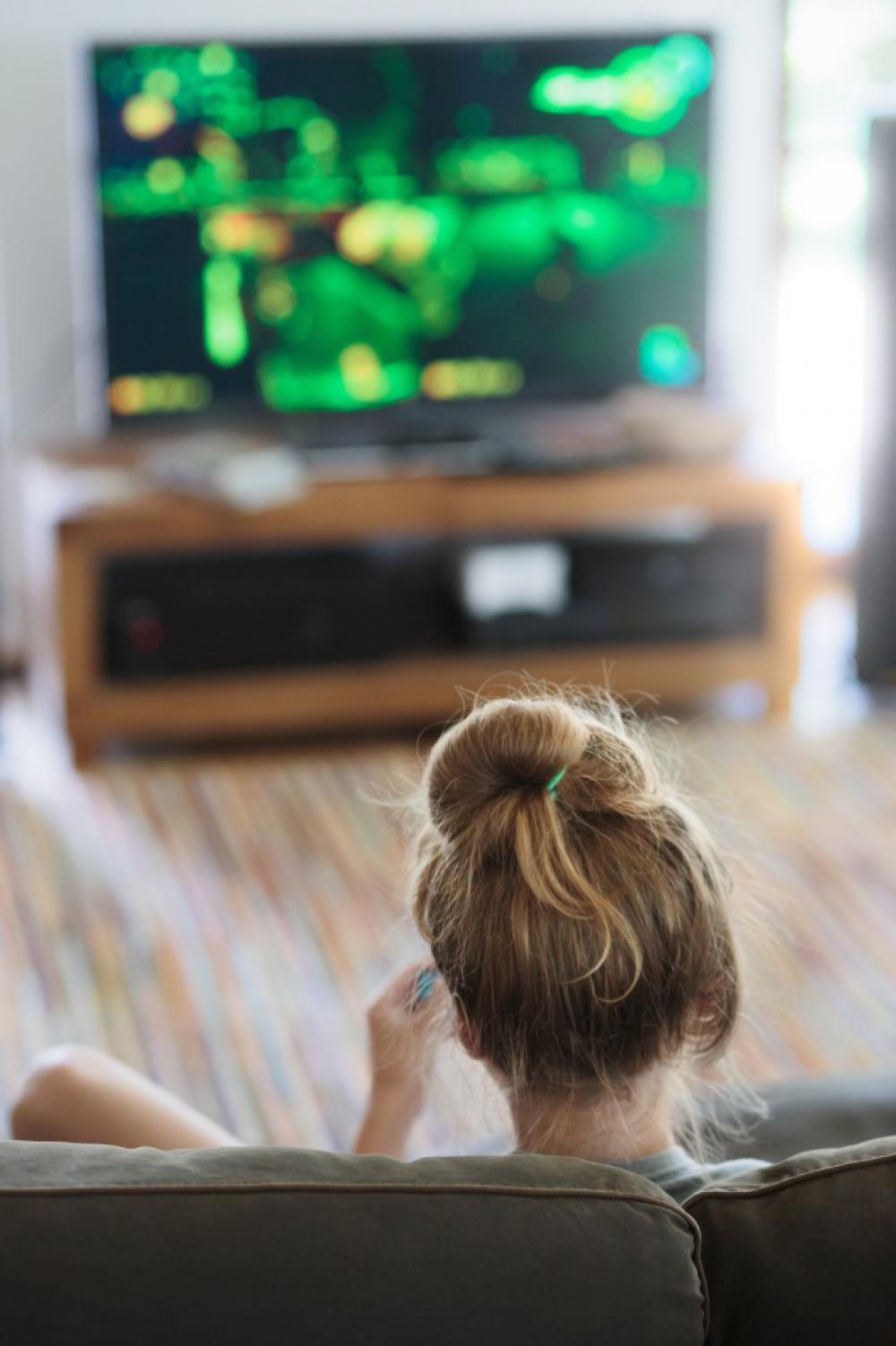 Back view of young blonde girl relaxing and playing video game sitting on sofa in front of tv screen at home Back view of young blonde girl relaxing and playing video game sitting on sofa in front of tv screen at home