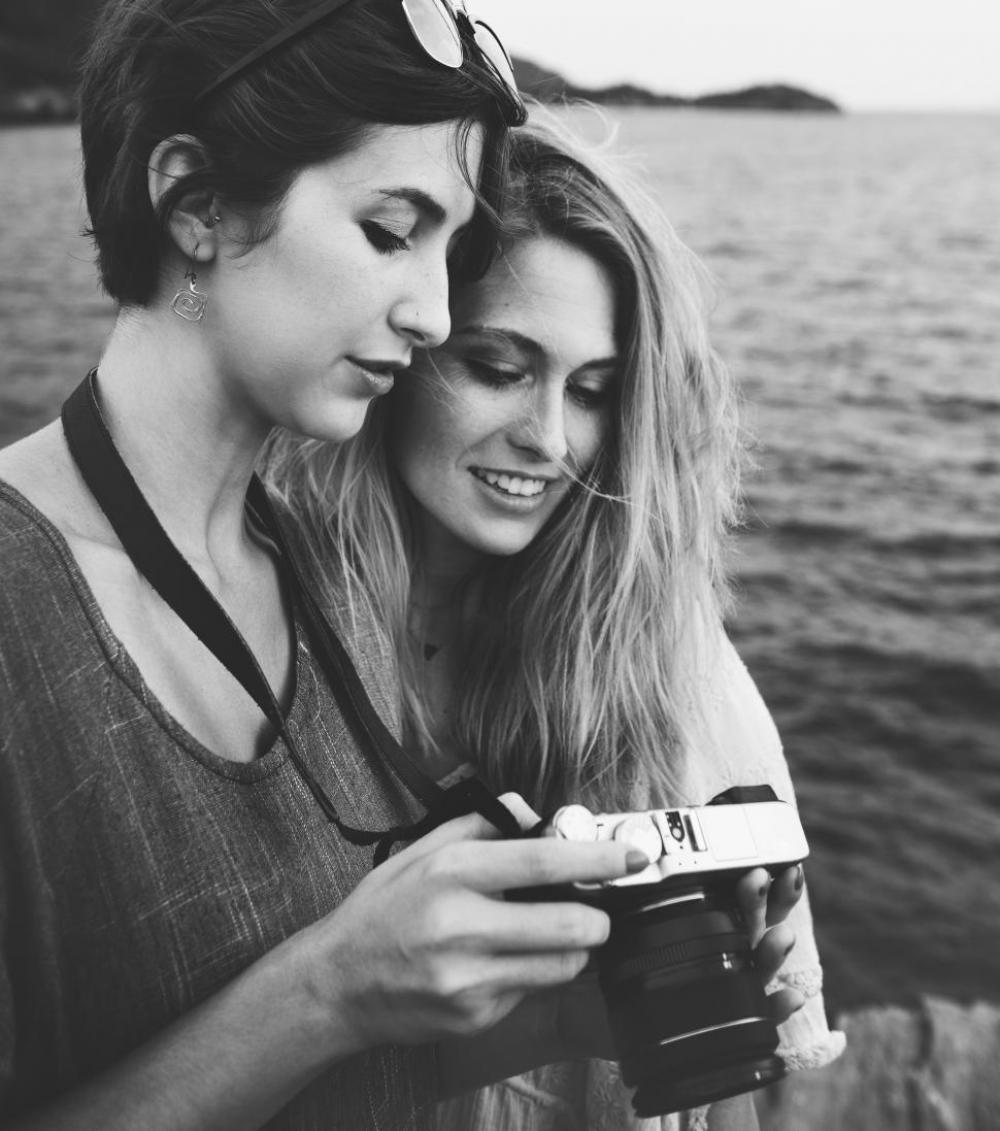 Two young caucasian women looking at their photograph on the camera display by the sea Two young caucasian women looking at their photograph on the camera display by the sea