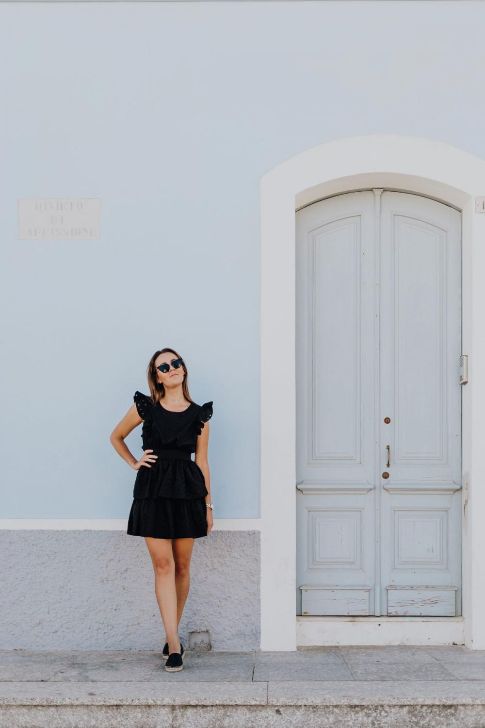 A young woman with dark hair wearing a black dress poses by the blue building A young woman with dark hair wearing a black dress poses by the blue building