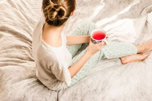 Attractive young girl in a pajamas holding a cup of tea while sitting in white bedding
