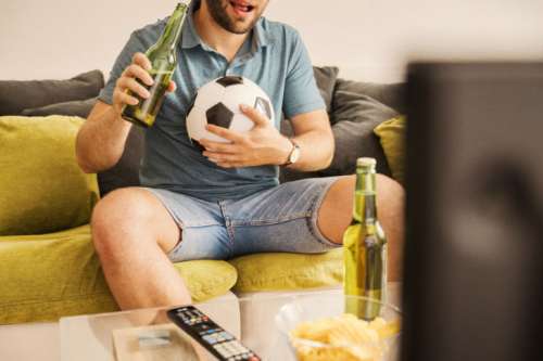 Young man watching football on TV and drinking beer at home