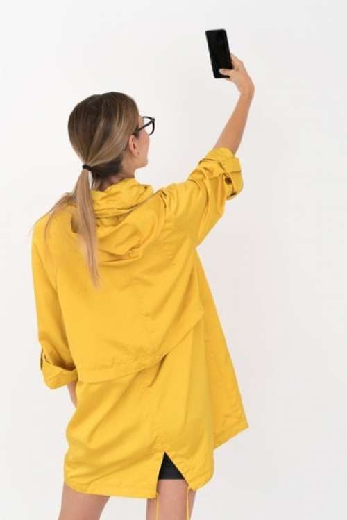 Young Blonde Woman In Yellow Raincoat And Black Glasses Taking A Selfie