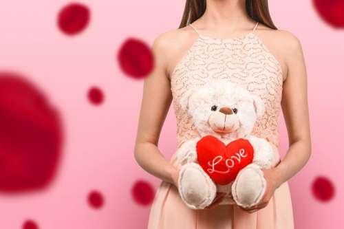 A young girl in a pink dress holding a teddy bear with a heart of love. Pink background. Blured rose flowers.