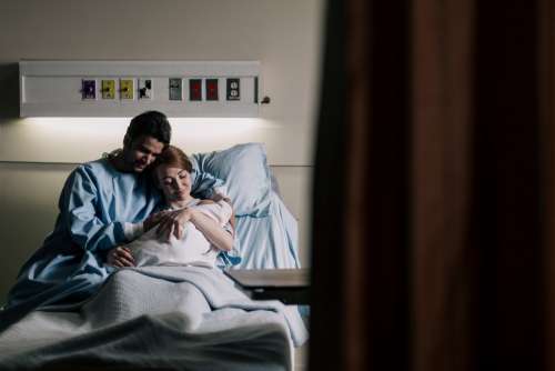 Mother And Father Embrace In Hospital Bed While Admiring Baby Photo