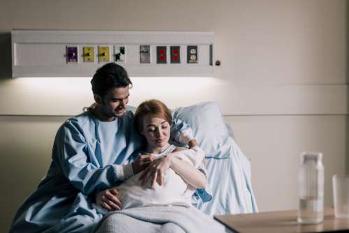 Mother And Father Sitting In Hospital Bed Admiring Newborn Photo