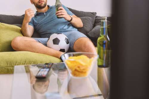 Young man watching football on TV and drinking beer at home