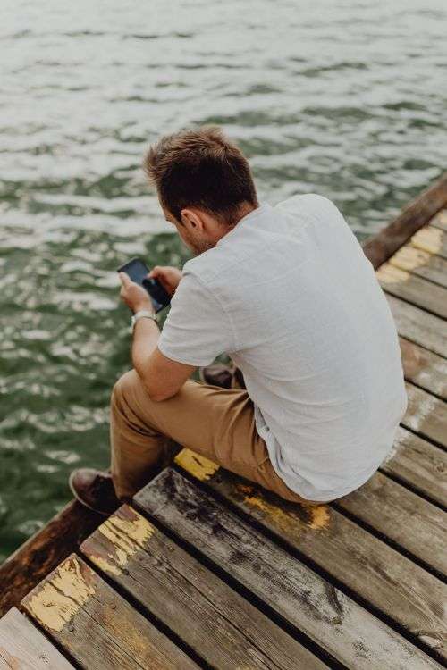 The man is using his phone at the lake