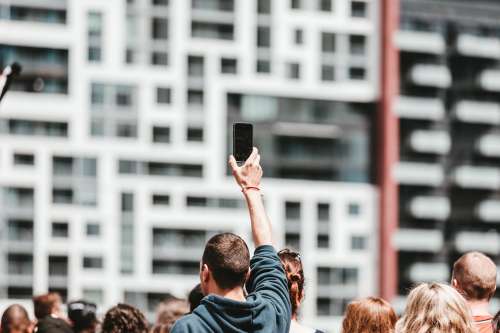A Man Holds Up A Mobile In A Crowd On A Bright Day Photo