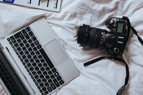 Silver laptop, a camera, magazines and other items on white bed sheets