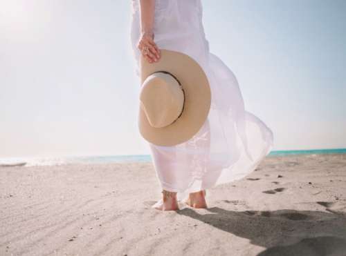 Portrait of beautiful woman in white dress and hat in hand walking on beach near sea or ocean 