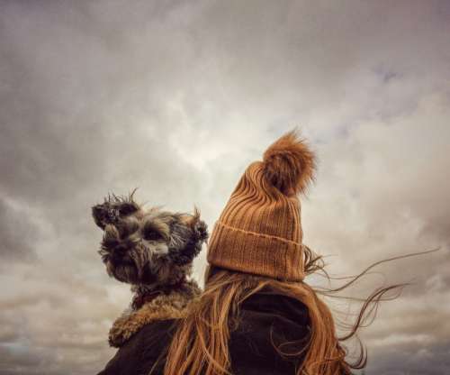 A rear view of fashionable young girl wearing a woolly bobble hat and holding her scruffy pet dog on her shoulder on a windy day in autumn or fall
