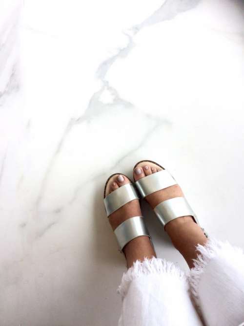 Bold metallic silver sandals worn by woman against white marble floor in minimalist setting. 