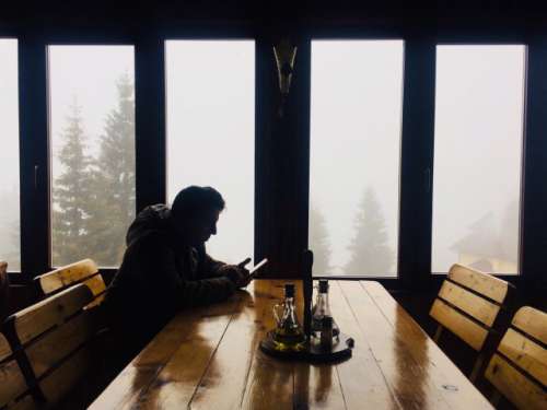 Single man in winter clothes texting at a table in a rustic restaurant with fog and trees seen out of the windows