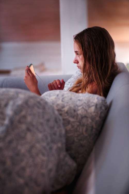 Teenage girl watching video online on smartphone sitting on sofa at home in the evening