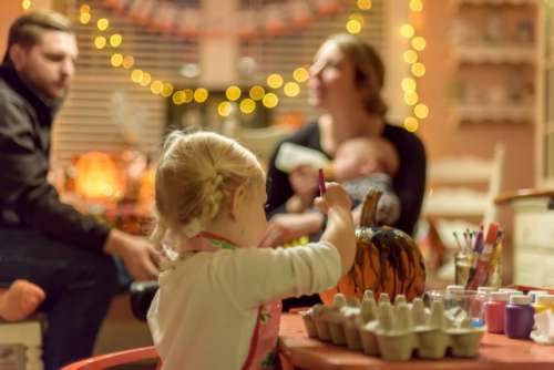 Little girl painting a pumpkin at a Halloween party while adults chat in the background

💫