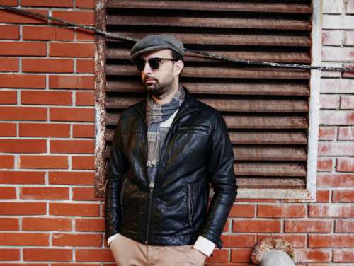 An urban portrait of a young man with a beard. He's wearing a bright sweater, a scarf, a leather jacket and a hat. He's standing in front of a brick wall and wearing sunglasses. Street photography, urban backdrop, fashion, person, man, young adult.