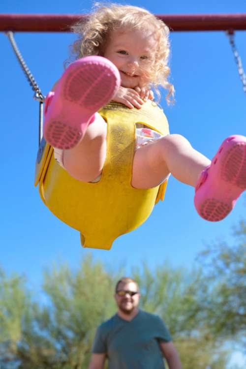 Toddler girl swinging high up in the air with Dad in background at playground
