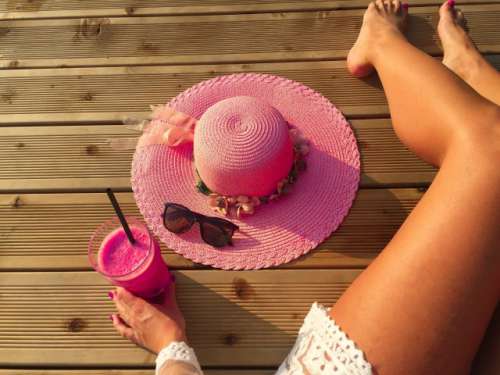 Woman's tanned sexy legs on wooden pontoon with glass of raspberry smoothie and pink summer hat 