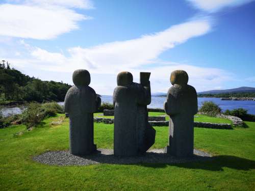 Three Stone Figures On A Grassy Knoll Overlook A River Photo