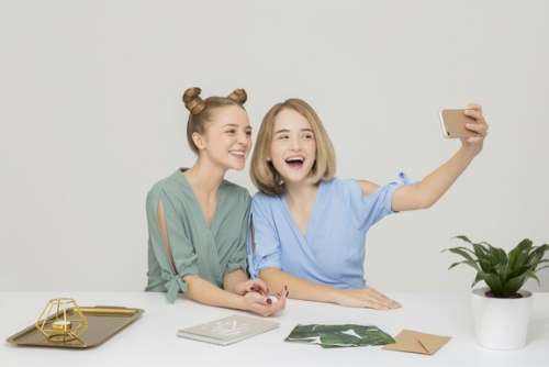 Two Smiling Girls Sitting At The Table And Making A Selfie