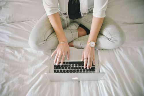 Woman’s Hands With Jewellery Typing On Laptop Sitting On A Bed