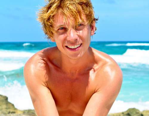 Tanned Blond Boy Showing Tattoo On A Beach