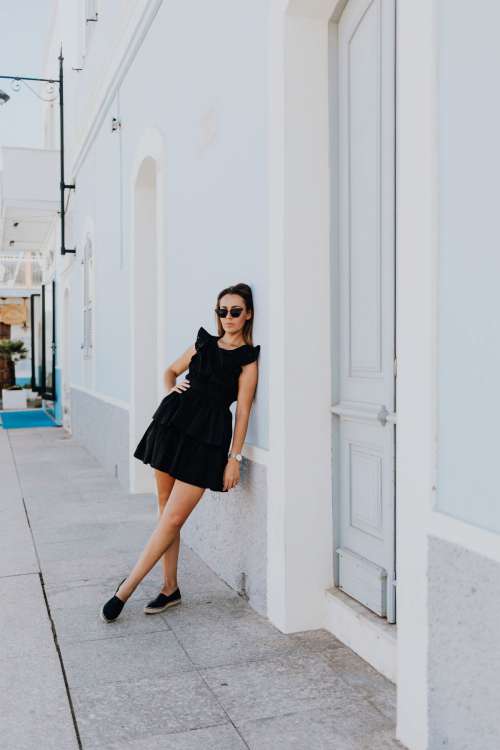 A young woman with dark hair wearing a black dress poses by the blue building