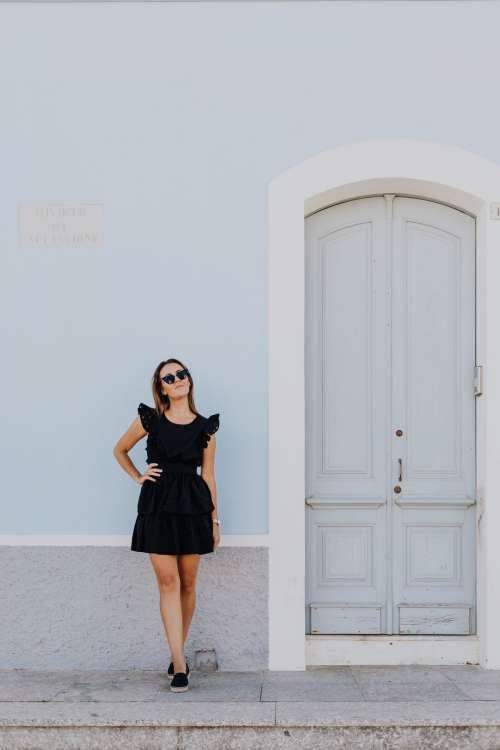 A young woman with dark hair wearing a black dress poses by the blue building