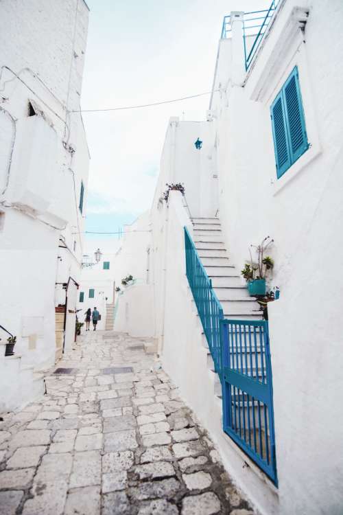 Two People Walking Around White Buildings And Blue Shutters Photo