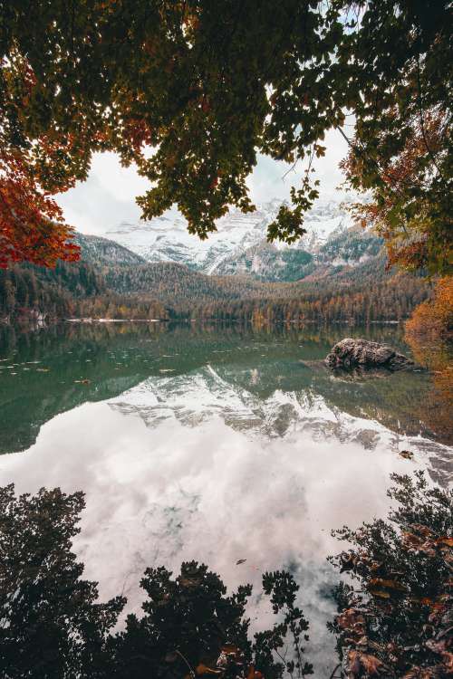 Snow Covered Mountains Reflect On The Water By A Fall Forest Photo