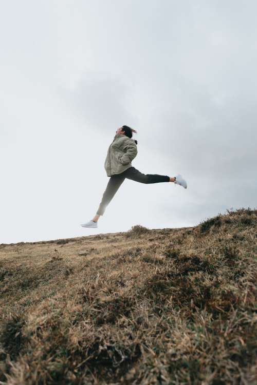 Person Jumps High With Legs Out Above A Brown Grassy Hill Photo
