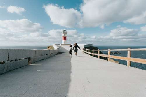 Two People Run Down A Paved Road Towards A Lighthouse Photo