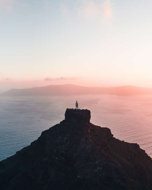Person Standing On A Far Cliff Taking A The Setting Sun Photo