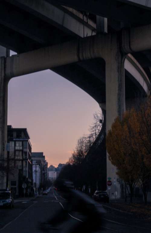 A Sun Sets Behind City Buildings From Under Bridge Photo
