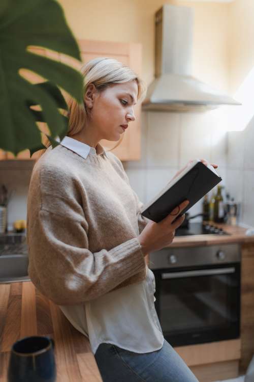 Woman Stands In Her Kitchen Reading A Book Photo
