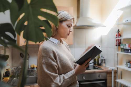 Woman Stands In Her Kitchen With A Book In Hand Photo