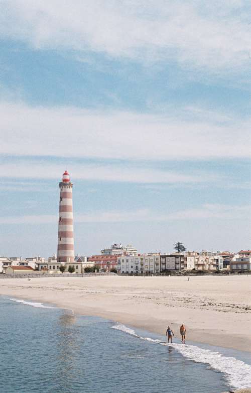 Two People Walk The Beach With A Lighthouse In The Distance Photo