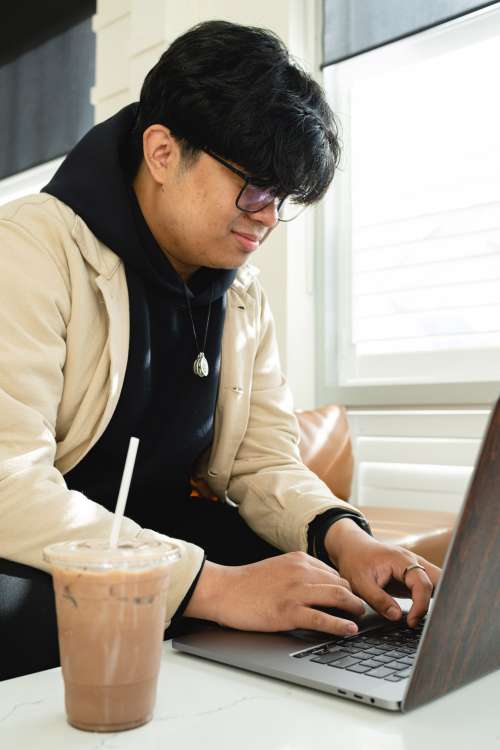 Man In Glasses Types On A Laptop Beside Iced Coffee Photo