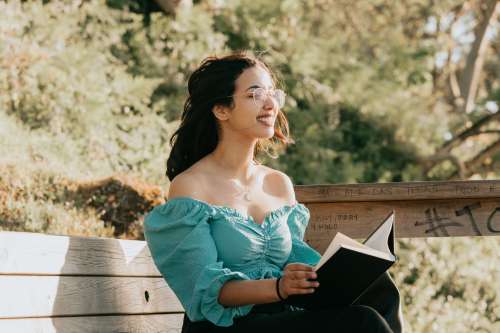 Woman Sits On A Bench Outdoors With A Book Smiling Photo