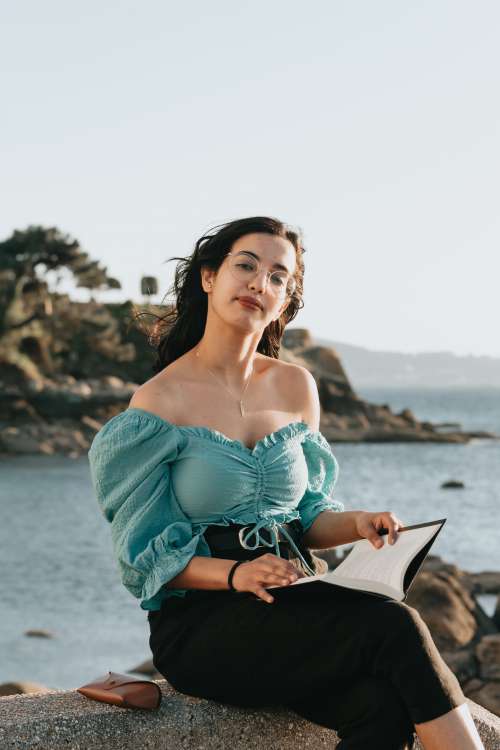 Woman Holds A Book And Sits On Rocks By The Water Photo