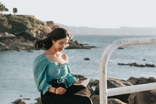 Woman Reads A Book With Rocks And Water Behind Her Photo
