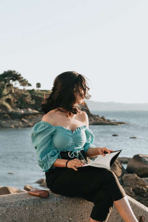 Woman Sits By The Water And Holds A Novel Open Photo