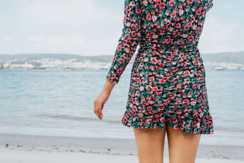 Person In A Floral Dress Stands On A Beach Facing The Water Photo