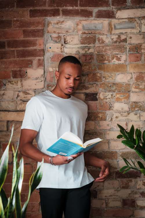Person In White Stands Reading A Novel By A Brick Wall Photo
