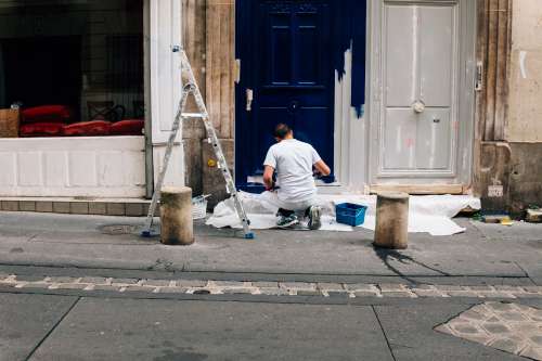 Person Crouches Down On A Street And Paints A Door White Photo