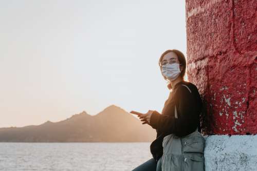 Woman In Facemask Leans Against A Wall By Water Photo