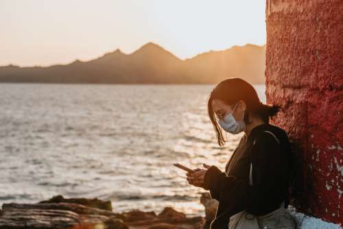 Woman In A Facemask Looks At Her Phone Standing By Water Photo