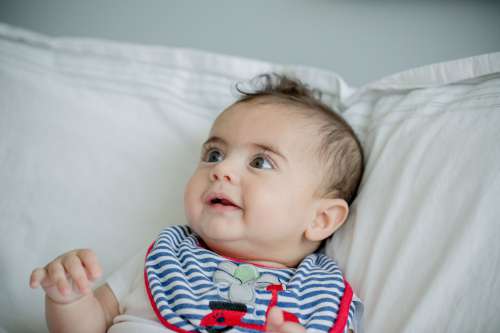 Smiling Baby Wearing A Bib Leans Against A White Pillow Photo