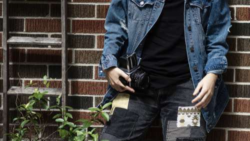 Person In Blue Jean Jacket Holds Camera Against Red Brick Wall Photo