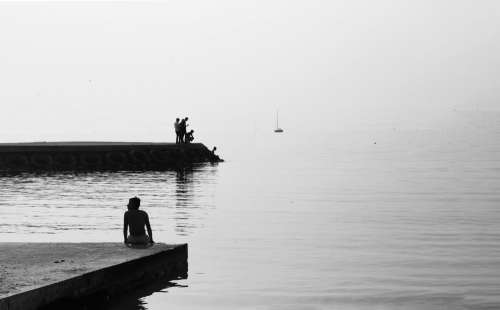 Black And White Photo Of People On A Cement Shore By Still Water Photo